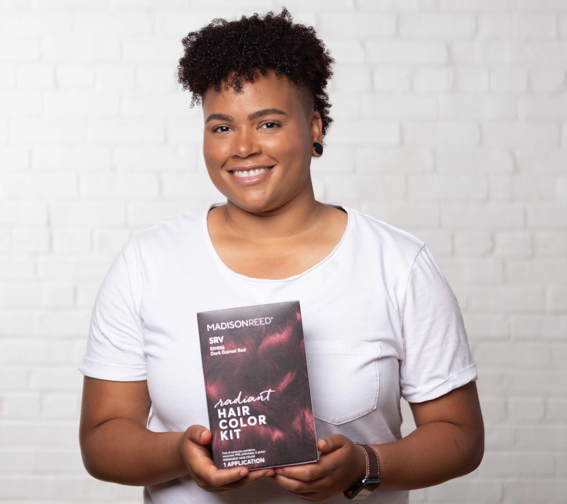 Woman with short coily hair smiling and holding Madison Reed Radiant Hair Color Kit